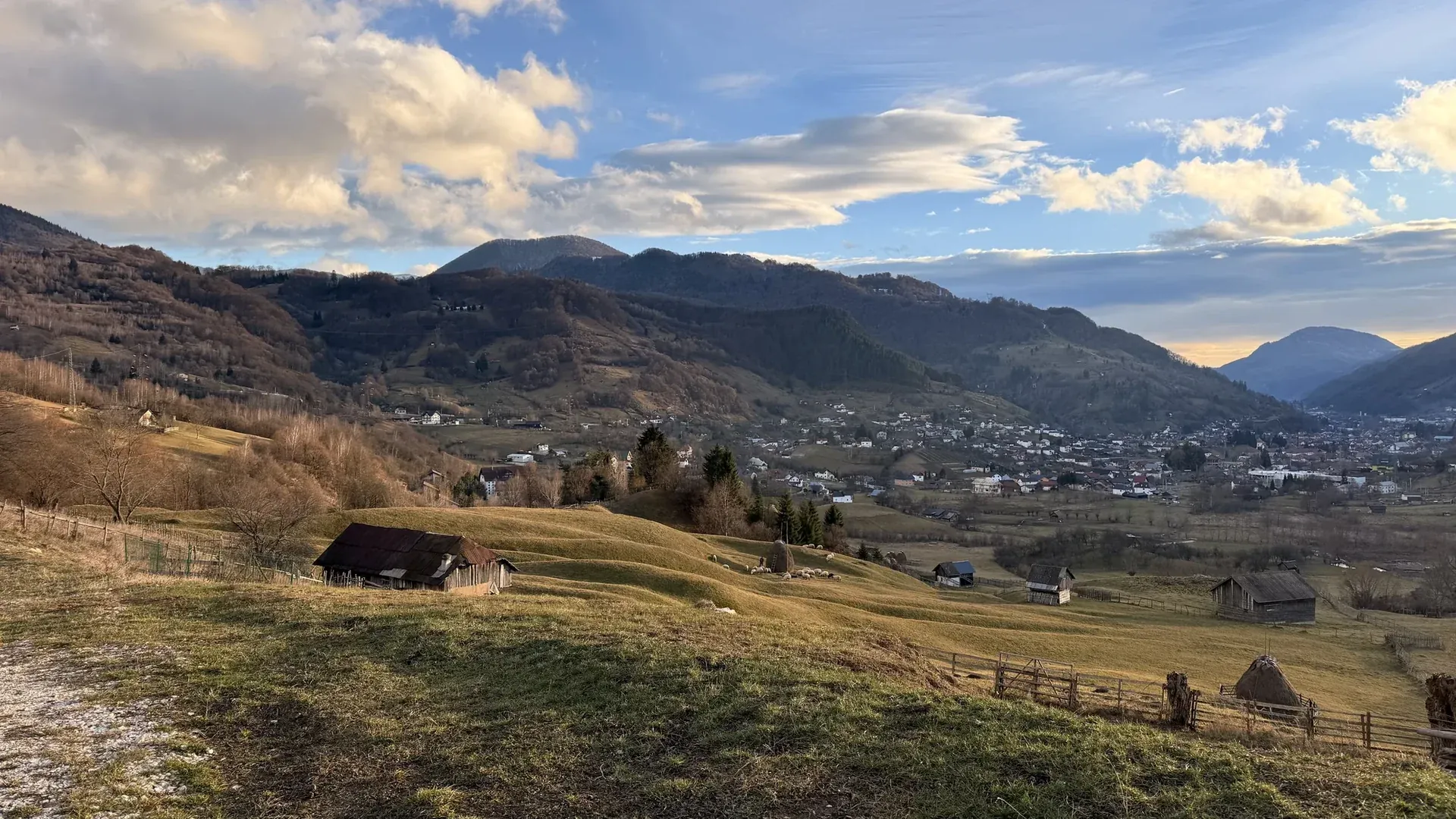 A pasture with a wooden hut and fence above the village, under an evening sky